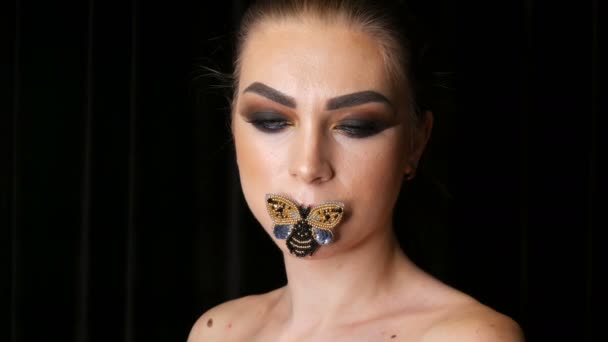 一个美丽的年轻女孩模型的肖像与时尚的烟熏眼睛化妆和蝴蝶在她的嘴唇摆姿势在镜头前在演播室。高时尚 — 图库视频影像