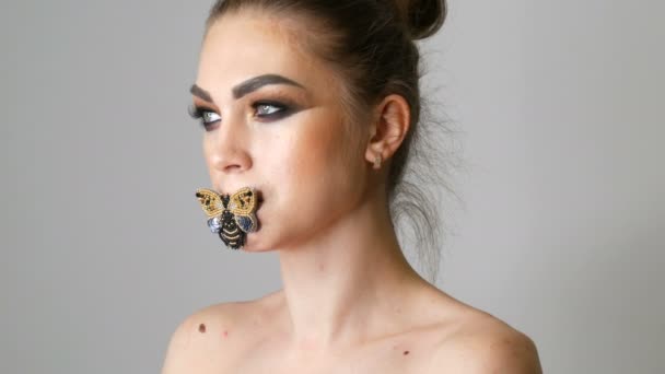 Πορτραίτο του μια όμορφη νεαρή κοπέλα μοντέλο με μακιγιάζ μόδας καπνιστή μάτια και μια πεταλούδα στα χείλη της, ποζάρει μπροστά από την κάμερα στο στούντιο. Υψηλός-μόδας — Αρχείο Βίντεο
