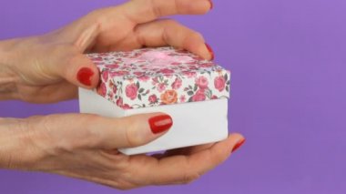 Bir çiçek baskı ve tırnakları mor bir arka plan üzerinde kırmızı oje ile boyanmış kadın elleri tarafından düzenlenen pembe bir yay ile küçük bir hediye kutusu açın