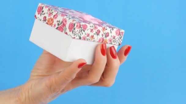 Uma pequena caixa de presente com uma impressão floral e um arco rosa que é realizada pelas mãos da mulher cujas unhas são pintadas com esmalte vermelho em um fundo azul — Vídeo de Stock