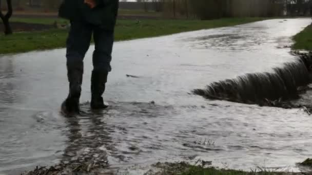 灾难。村子里洪水泛滥。肮脏的黑河沿着道路流淌。穿着黑色橡胶靴的男人穿过水坑. — 图库视频影像