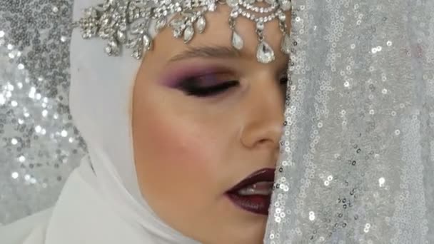 Modèle professionnel dans une image blanche et une tiare argentée avec un maquillage multicolore lumineux appelé un œil fumé, posant devant la caméra en agence de mannequins sur un fond argenté. Haute couture — Video