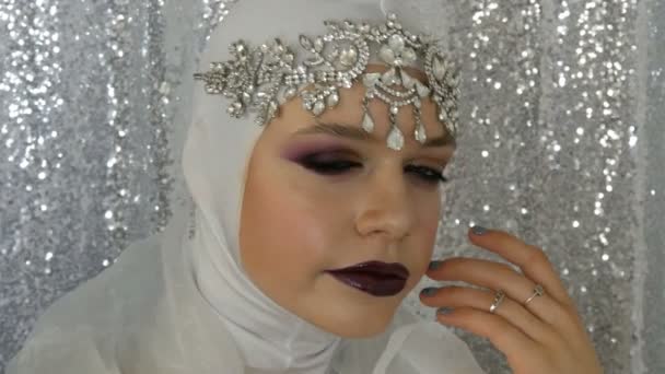 Visage du modèle dans une image blanche et une tiare argentée avec un maquillage multicolore lumineux appelé un œil fumé, posant devant la caméra en agence de mannequins sur un fond argenté. Haute couture — Video