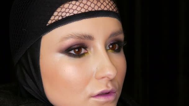 Professionell tjej modell med vacker makeup utgör i en svart mössa på huvudet framför kameran på svart bakgrund i bilden av en svart änka. Hög modegrad — Stockvideo