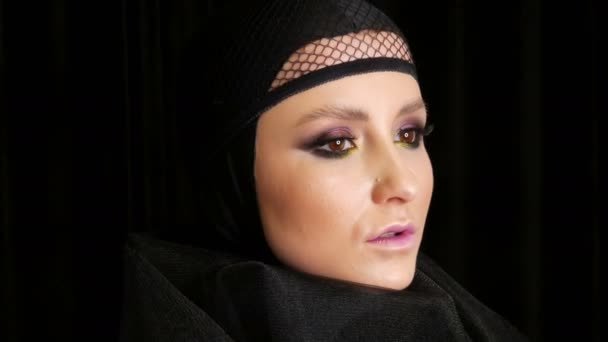 Profi-Model mit wunderschönem Make-up posiert in einer schwarzen Mütze auf dem Kopf vor der Kamera auf schwarzem Hintergrund in dem Bild einer schwarzen Witwe. Mode im Trend — Stockvideo