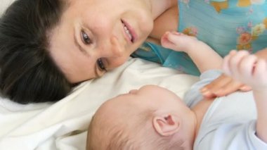 Uzun koyu saçlı genç güzel anne beyaz bir yatakta yatıyor ve iki aylık yeni doğan bebek ile oynuyor