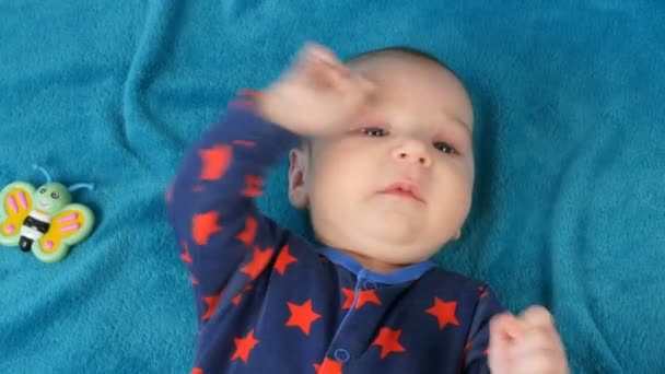 Porträt eines lustigen kleinen Neugeborenen von zwei Monaten, das auf einer blauen Bettdecke liegt, umgeben von Babyrasseln — Stockvideo