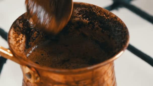 Чайная ложка смешивает черный молотый кофе с медным турком, который стоит на белой газовой плите — стоковое видео