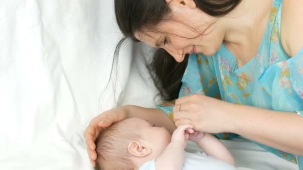 Молода красива мати з довгим темним волоссям лежить на білому ліжку і грає з новонародженою дитиною два місяці — стокове відео