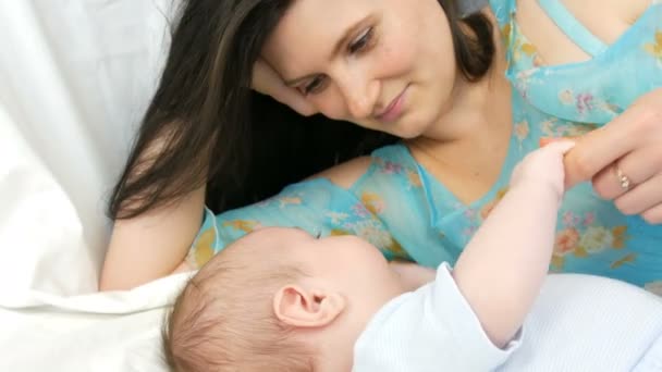 Молодая красивая мать с длинными темными волосами лежит на белой кровати и играет с новорожденным ребенком двух месяцев — стоковое видео