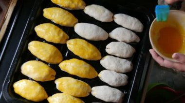 Fırında pişirilmeye hazır, bir sıra uzun hamur topları fırın tepsisi üzerinde. Kadın eli çırpılmış yumurtayı özel mutfak silikon fırçasıyla yağlar.