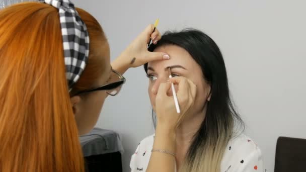 Make-up kunstenaar met rood haar en een bril die professionele make-up doet. Mooie avond make-up wordt toegepast op een jonge vrouw — Stockvideo