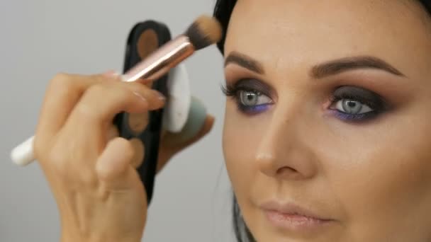 Make-up kunstenaar leidt een vrouw in het gezicht met een borstel voor het aanbrengen van poeder, stichting of concealer. Lilac en parelmoer rokerige ogen oogschaduw, ogen en gezicht van een vrouw close-up. Professionele high fashion. — Stockvideo