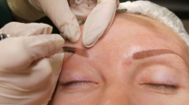 Beyaz eldivenli bir güzellik uzmanının elleri özel kalem yardımıyla tenine pigment sürer. Kaş şekli düzeltmesi. Mikro patlayıcı, kalıcı makyaj, kalıcı makyaj, barut püskürtme.