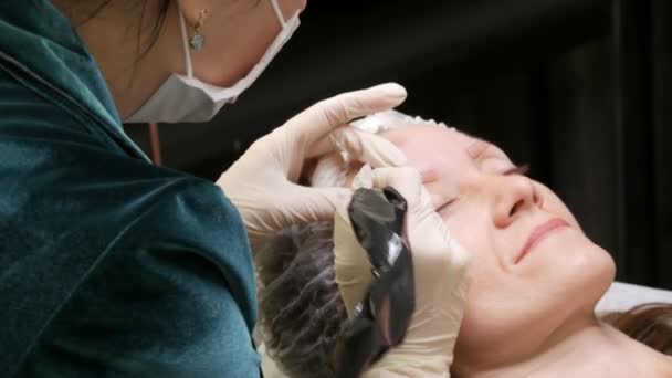 Um esteticista de máscara com casaco verde e luvas brancas aplica pigmento na pele com a ajuda de uma caneta especial. Correção da forma da sobrancelha. Microblading, tatuagem, pulverização em pó, maquiagem permanente — Vídeo de Stock