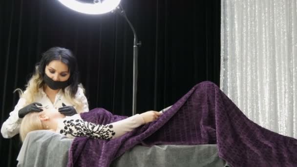Gyönyörű nő stylist sminkes művész kozmetológus teszi szemöldök korrekció a beteg arcát, amely fekszik egy speciális kanapén elegáns szépség stúdió fekete-ezüst függönyök