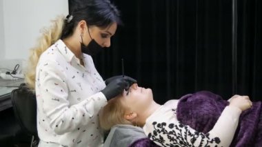 Güzellik kliniğinde kozmetoloji prosedürü. Kaşların şeklini modellemede usta olan güzel kadın, koltukta yatan müşterisine kaş düzeltmesi yapıyor. Mikropatinaj, dövme, kalıcı makyaj.