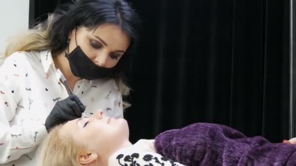 Hermosa mujer estilista maquillaje artista cosmetólogo hace corrección de cejas en la cara del paciente que está acostado en un sofá especial en elegante estudio de belleza con cortinas negras y plateadas — Vídeo de stock