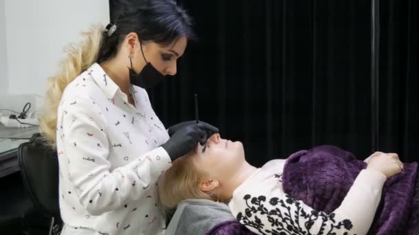 Procedimiento de cosmetología en una clínica de belleza. Hermoso maestro en el modelado de la forma de las cejas hace corrección de cejas a su cliente, que se encuentra en el sofá. Microblading, tatuaje, maquillaje permanente — Vídeos de Stock