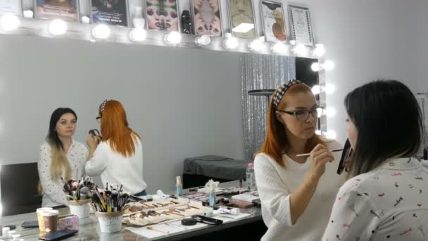 Szeptember 20, 2019 - Kamenskoye, Ukrajna: Stílusos nő make-up művész vörös hajjal alkalmazó professzionális smink modellek arc szépségszalon — Stock videók