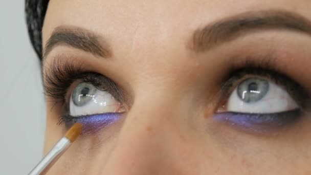 Krásné modré oko pro mladou ženu, která s pomocí speciálního štětce nanáší kouřové oči Lilac a perleťově zakouřené oči, oči a tvář ženy zblízka. Profesionální high fashion. — Stock video
