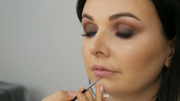 Visage incroyablement beau d'une jeune femme aux yeux bleus, maquillage professionnel aux tons lilas, yeux fumés, maquilleuse applique doucement du rouge à lèvres rose sur ses lèvres — Video