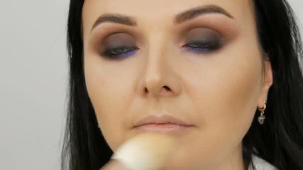 Maquillaje artista lleva a una mujer en la cara con un cepillo para la aplicación de polvo, fundación o corrector. Ojos ahumados lila y nacarada sombra de ojos, ojos y cara de una mujer de cerca. Profesional de alta moda . — Vídeo de stock