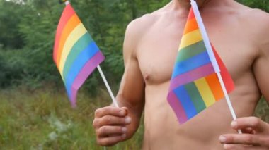 Gövde, ellerinde gökkuşağı, ellerinde yeşil doğanın arka planına karşı alışılmadık oryantasyonun sembolü olan Gay gururu LGBT bayrağını tutan bronz tenli genç bir adamın atletik vücudu.