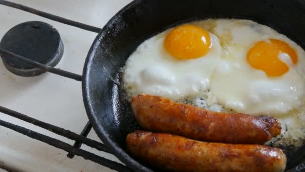 Funny breakfast in the pan in the form of a face and a smile with eyes. Two fried eggs and thick Bavarian sausages fried in a vintage cast iron skillet on a gas stove — Stock Video