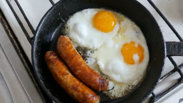 Αστείο πρωινό στο τηγάνι με τη μορφή ενός προσώπου και ένα χαμόγελο με τα μάτια. Δύο τηγανητά αυγά και χοντρά βαυαρικά λουκάνικα τηγανισμένα σε ένα παλιό τηγάνι από χυτοσίδηρο σε μια κουζίνα γκαζιού. — Αρχείο Βίντεο