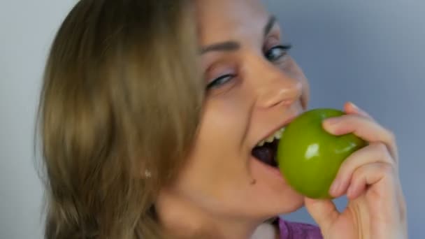 Gezicht portret van een jonge mooie vrouw met een eetlust en grappig eten van een grote groene appel, afbijten stukken. Vegetarisme concept, vers fruit in de handen van een jong meisje, gezond voedsel — Stockvideo