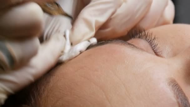 Beautician en guantes negros hace la corrección permanente del maquillaje de la forma de las cejas a una muchacha hermosa joven. Microblading, tatuaje de la ceja en un salón de belleza — Vídeos de Stock