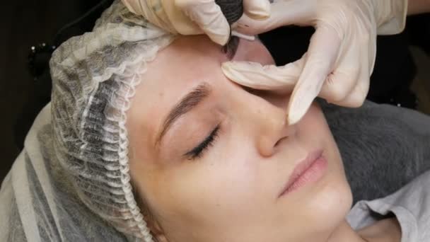 Una máquina especial del tatuaje de la aguja hace la corrección permanente del maquillaje de una ceja joven de las mujeres. Se inyecta un pigmento de pintura oscura debajo de la piel. Microblading, pulverización en polvo — Vídeos de Stock