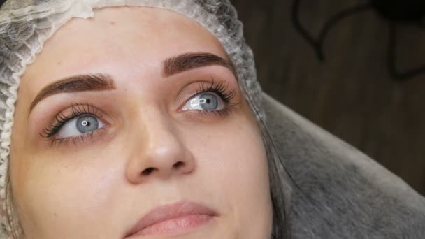 El retrato de la cara de una hermosa mujer joven con ojos azules y pestañas largas después del procedimiento de maquillaje permanente por microblading con tatuaje de cejas se encuentra en un sofá en un salón de belleza c — Vídeo de stock