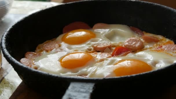 Smaczne, świeże jajka smażone na patelni z żeliwa, z przyprawami, pomidorami, słodką papryką i kiełbasą, z parą wodną. Poranne śniadanie, okrągłe żółtka jaj w domowej kuchni — Wideo stockowe