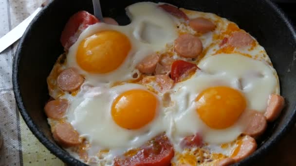 A man eats with a fork a delicious fresh fried egg in a vintage cast-iron frying pan, with seasonings, tomatoes, sweet paprika and sausages, with steam. Morning breakfast in home kitchen — Stock Video