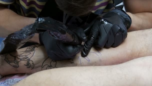 Van dichtbij gezien maakt een professional een tatoeage op het been van een man in een salon. Tekening met draaibaar tattoo machinegeweer — Stockvideo