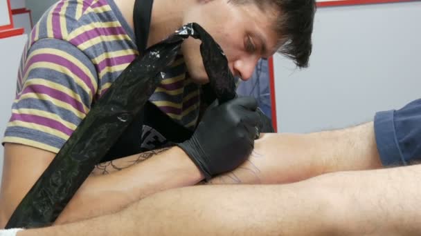 Retrato de um artista moderno enquanto trabalha com um cliente. Um profissional faz uma tatuagem na perna de um homem numa sala de estar. Desenho com metralhadora de tatuagem rotativa — Vídeo de Stock