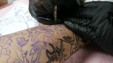 Bir profesyonelin yakından bakınca, salondaki bir adamın bacağına dövme yapıyor. Döner dövmeli makineli tüfekle çizim