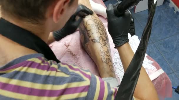 Retrato de un artista moderno mientras trabaja con un cliente. Un profesional hace un tatuaje en la pierna de un hombre en un salón. Dibujo con ametralladora rotativa del tatuaje — Vídeo de stock