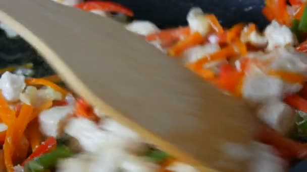 Азиатская еда. Овощи жарятся на сковороде с кусками курицы. Паприка, морковь, лук, чеснок, зеленые стручки, смешанные с деревянной кухонной лопаткой и тушеным — стоковое видео