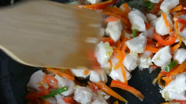 Азиатская еда. Овощи жарятся на сковороде с кусками курицы. Паприка, морковь, лук, чеснок, зеленые стручки, смешанные с деревянной кухонной лопаткой и тушеным — стоковое видео
