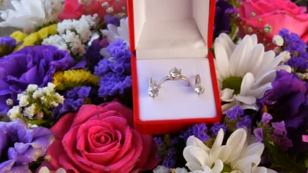 Cadeau aanwezig rode doos met een set van verlovingsring en oorbellen op de achtergrond van een veelkleurig mooi boeket bloemen. Huwelijk aanzoek. Witgoud of zilver met diamanten steen. — Stockvideo