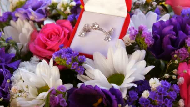 Ajándék jelen piros doboz egy sor eljegyzési gyűrű és fülbevaló a háttérben egy sokszínű gyönyörű csokor virág. Házassági ajánlat. Fehér arany vagy ezüst gyémántkővel. — Stock videók