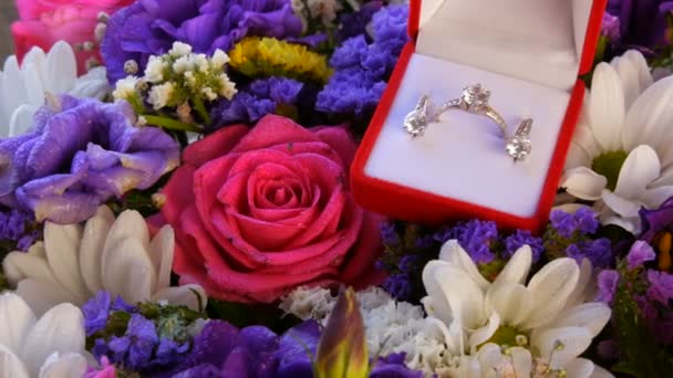 Cadeau aanwezig rode doos met een set van verlovingsring en oorbellen op de achtergrond van een veelkleurig mooi boeket bloemen. Huwelijk aanzoek. Witgoud of zilver met diamanten steen. — Stockvideo