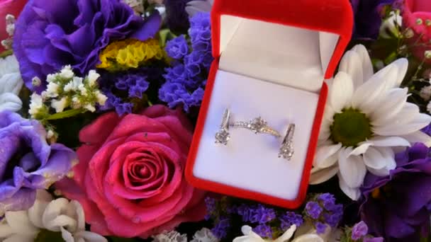 Geschenk Geschenk-Geschenk rote Box mit einem Satz Verlobungsring und Ohrringe auf dem Hintergrund eines bunten schönen Blumenstrauß. Heiratsantrag. Weißgold oder Silber mit Diamantstein. — Stockvideo