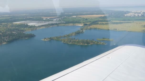 Bir yolcu uçağının beyaz kanadı, güzel mavi bir gölün doğa manzarasına karşı, bir göletin üzerinde uçar, yeşil ormanlar ve küçük evler ve kasabalar. Seyahat kavramı — Stok video