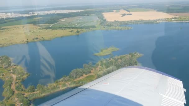 Біле крило пасажирського літака на тлі красивого блакитного озера, летить над ставком, зеленими лісами і маленькими будинками і містечками. Концепція подорожей — стокове відео