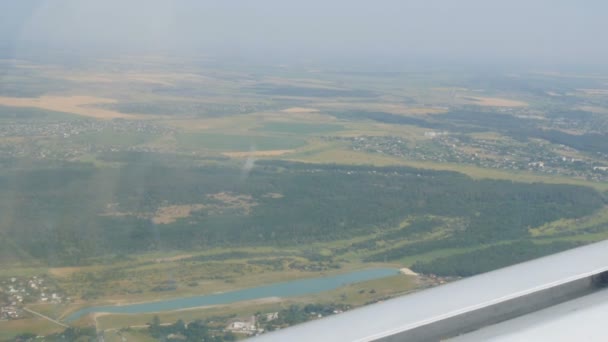 De witte vleugel van een passagiersvliegtuig tegen de top uitzicht op de natuur achtergrond van een prachtig blauw meer, vliegt over een vijver, groene bossen en kleine huizen en steden. Reisconcept — Stockvideo
