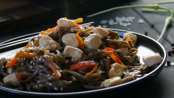 Asian buckwheat noodles soba with vegetables onion pods, carrots, paprika and sesame seeds and pieces of chicken, on a stylish black plate and Chinese chopsticks in the style of low key — Stock Video